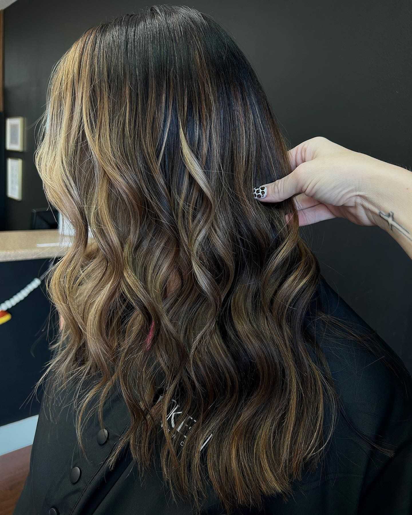 Woman with wavy, highlighted hair in a salon. Stylist's hand adjusts hair for a photo.